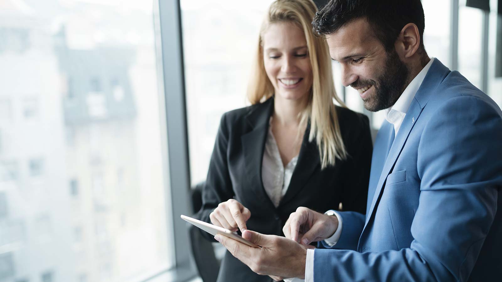 A brunette man and a blonde woman in suits smiling looking at a tablet together